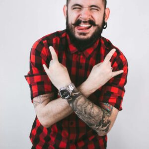 A stylish man in red plaid shirt with tattoos poses playfully indoors.